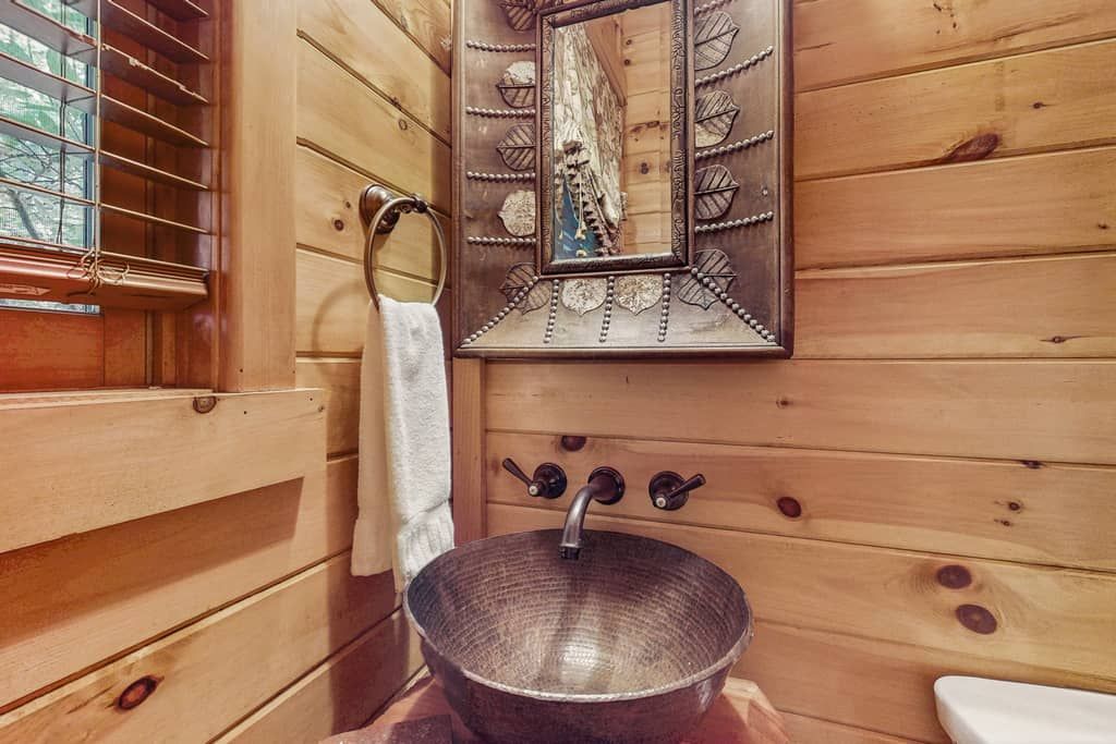 Ein Badezimmer einer Holzhütte mit einem Metallwaschbecken, einem Handtuch an einem Ring und einem Spiegel mit Blattmuster.