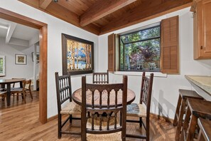 A dining area with a round wooden table and chairs, wooden ceiling beams, and a window with shutters. - Happy Hideaway on the West Shore by Tahoe Getaways - 4BR + Private Beach Access Near Tahoe City (Tahoe City)