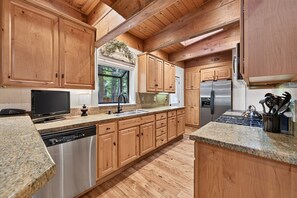 A kitchen with wooden cabinets, granite countertops, stainless steel appliances, and a window with a view of greenery. - Happy Hideaway on the West Shore by Tahoe Getaways - 4BR + Private Beach Access Near Tahoe City (Tahoe City)