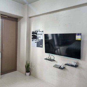 Ein Zimmer mit einem großen Flachbildfernseher an der Wand, einer Holztür, einem Blumentopf und einem an der Wand befestigten Regal mit elektronischen Geräten.