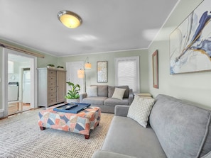 A living room with a sofa, ottoman, and a coffee table. There is a door leading to a kitchen, and a window with blinds.