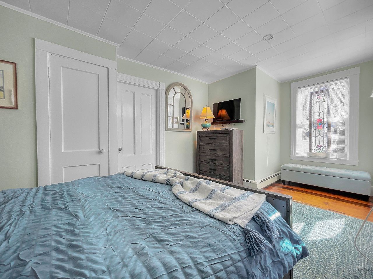 Een slaapkamer met een bed, een dressoir, een spiegel, een lamp en een raam.