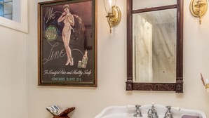 Un baño con inodoro blanco, lavamanos pedestal, un espejo grande y un anuncio antiguo en la pared.
