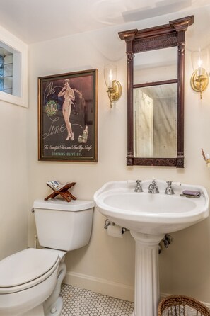 Une salle de bain avec des toilettes blanches, un lavabo sur colonne, un grand miroir et une affiche publicitaire d’époque accrochée au mur.