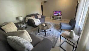 Ein modernes Wohnzimmer mit einem grauen Sofa, einem Couchtisch und einem Fernseher auf einem Standfuß.