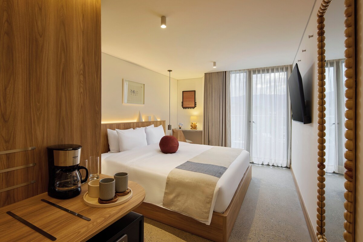 Habitación de hotel con cama, mesita de noche, cafetera y vistas al exterior a través de la ventana.