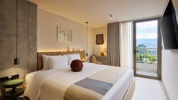 A hotel room with a large bed, a bedside table with a lamp, a view of the outdoors, and a flat-screen TV mounted on the wall.