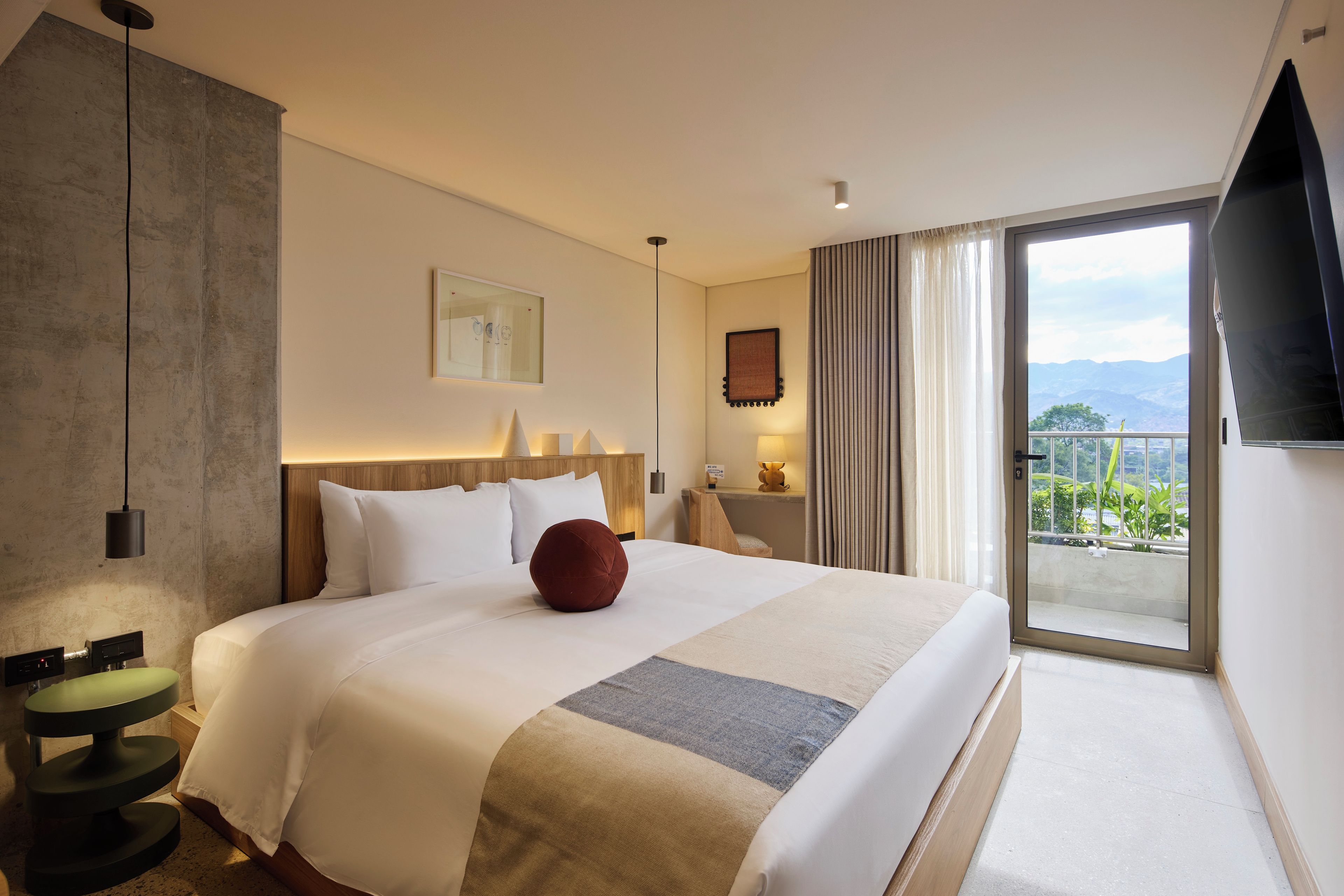 Habitación de hotel con una cama grande, una mesita de noche con lámpara, vistas al exterior y un televisor de pantalla plana montado en la pared.