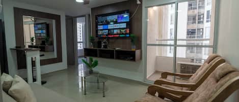 A modern living room with a flat-screen TV mounted on the wall, a ceiling fan, and a large window offering a view of the outside.