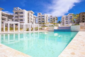 A swimming pool with a clear blue water surface, surrounded by modern buildings with balconies, and a clear sky above. - Stylish New 2-Bedroom Apartment with Pools & Gym (Cabarete)