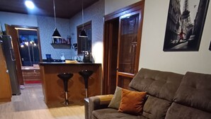 Una sala de estar con un sofá marrón, piso de madera y una cocina al fondo.