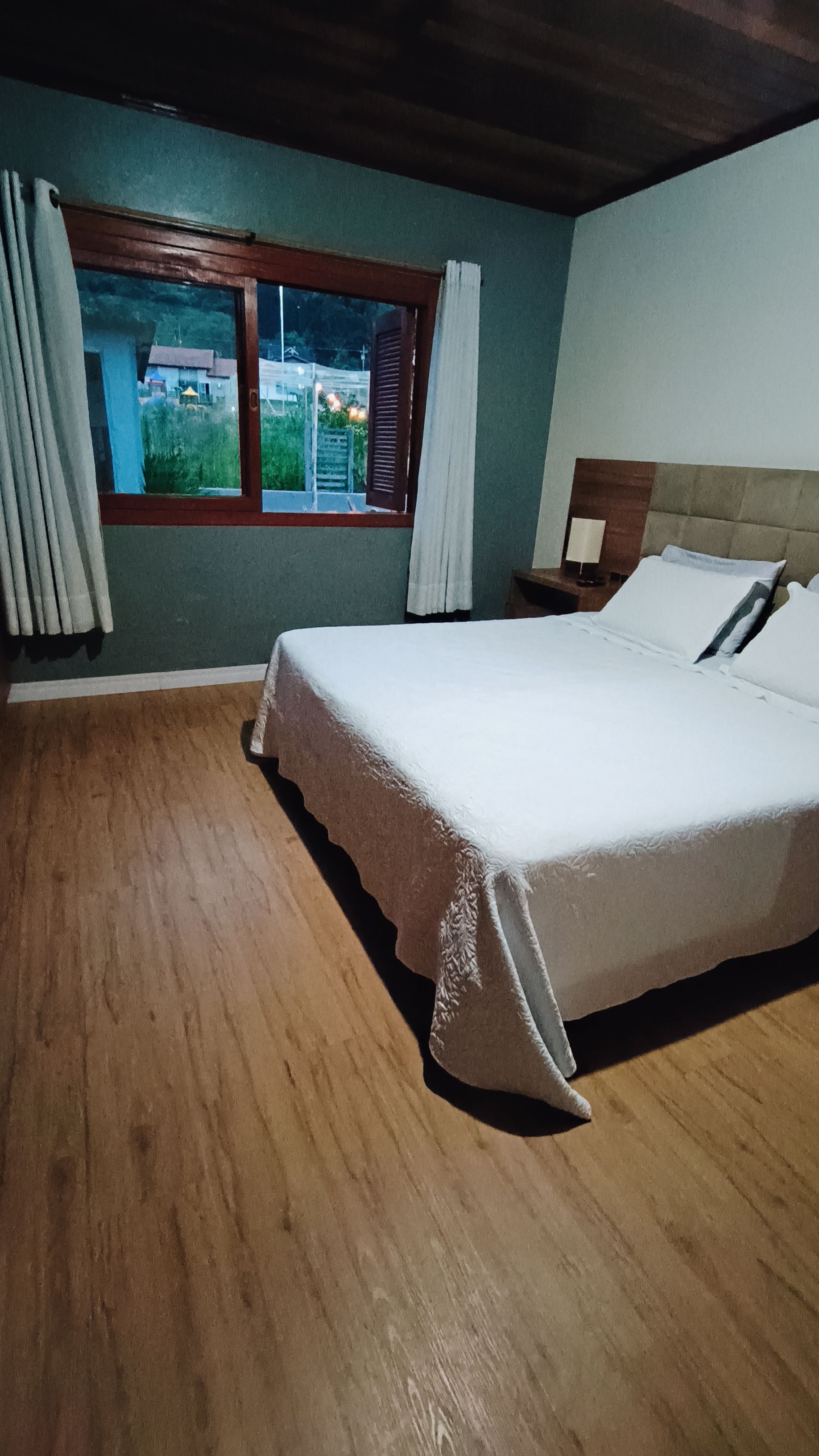 A bedroom with a wooden floor, a bed with white linens, a nightstand, a window with curtains, and a view of greenery outside.