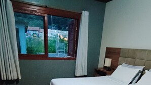 Un dormitorio con piso de madera, una cama con ropa blanca, una mesita de noche, una ventana con cortinas y vistas a zonas verdes.