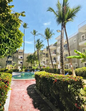 Un área de piscina con un sendero rojo, rodeada de palmeras y un edificio de varios pisos con balcones.