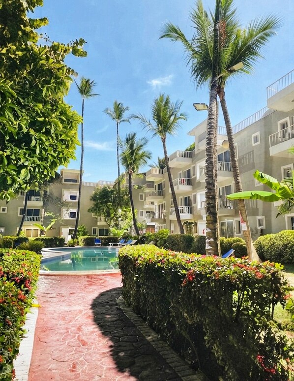 A pool area with a pathway leading to a building with balconies, surrounded by palm trees and greenery. - Gran sol caribe corales punta cana (Punta Cana)
