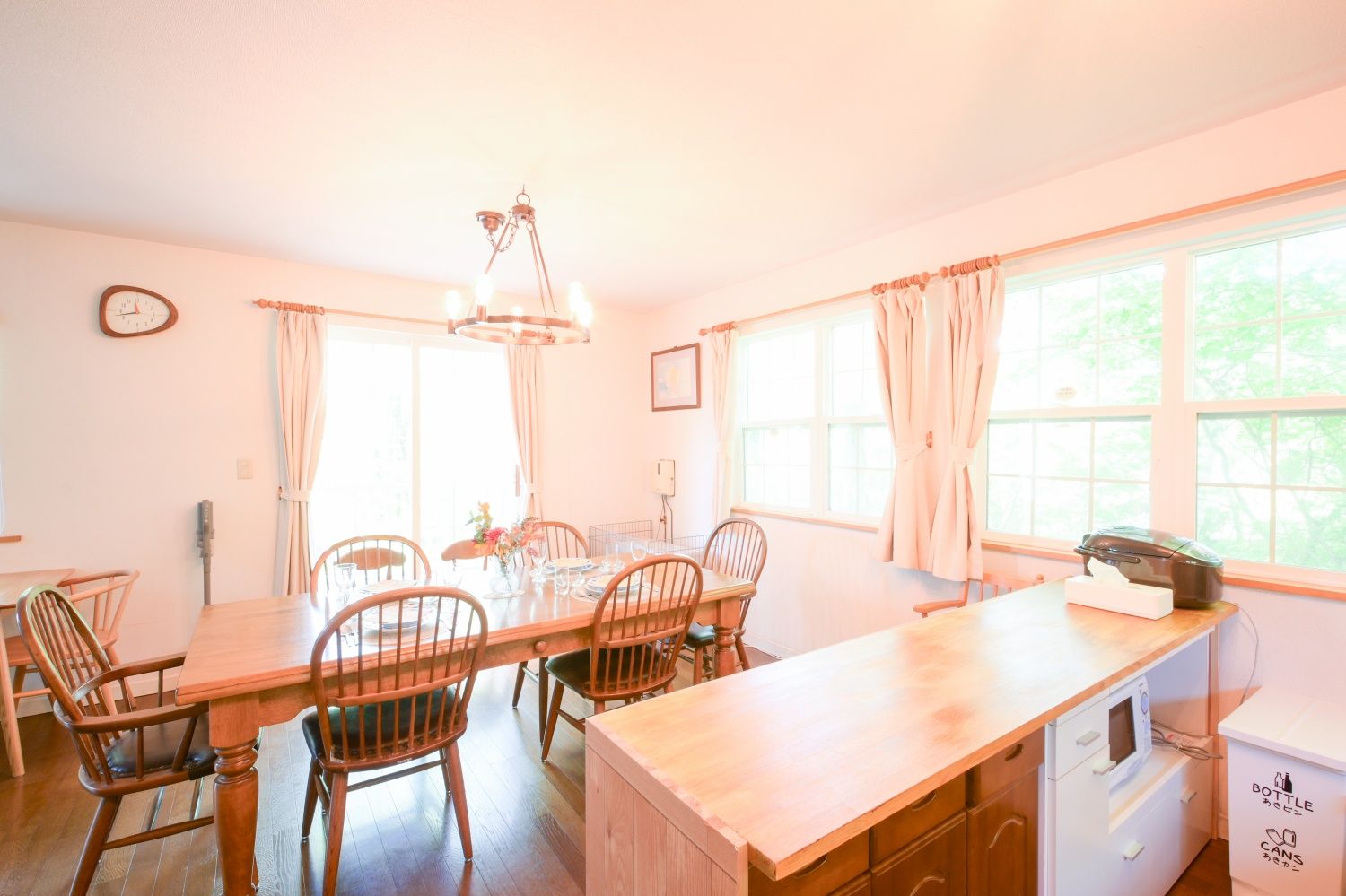 A dining area with a wooden table and chairs, a chandelier, and a kitchen area with a microwave and a refrigerator.