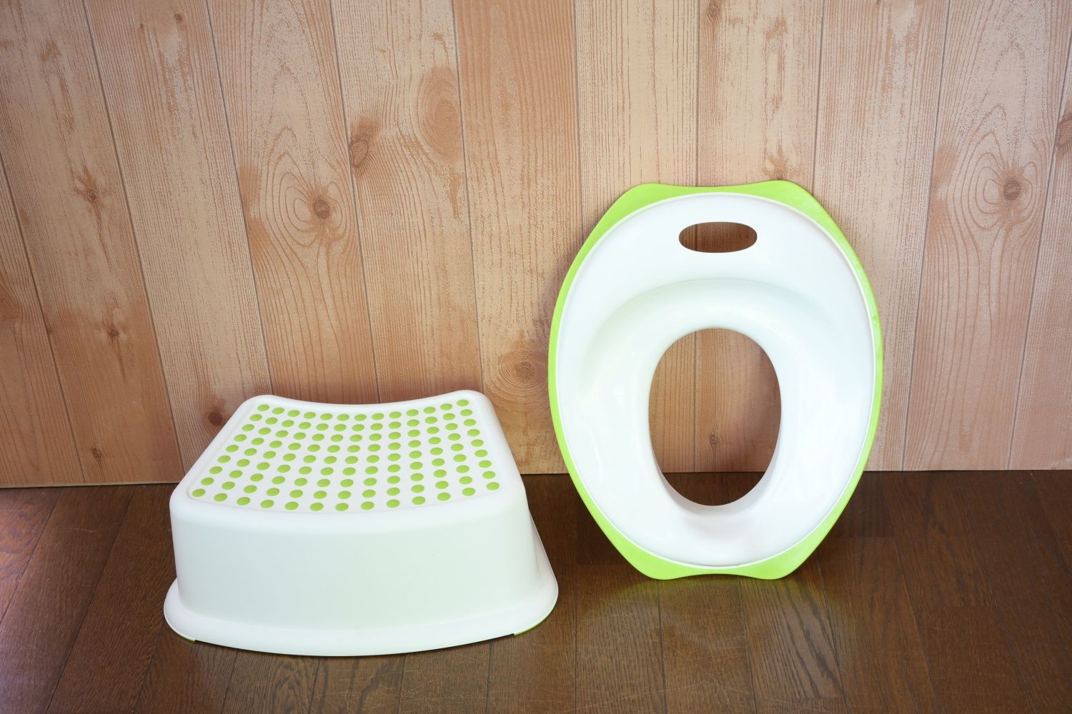 Un pot de bébé blanc et vert, doté d’un marchepied et d’un trou pour l’assise, est posé sur un plancher en bois, contre un mur également en bois.
