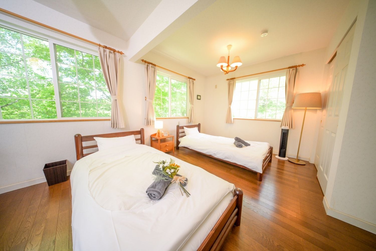 A hotel room with two beds, a wooden floor, a window with curtains, a bedside lamp, and a small trash bin.