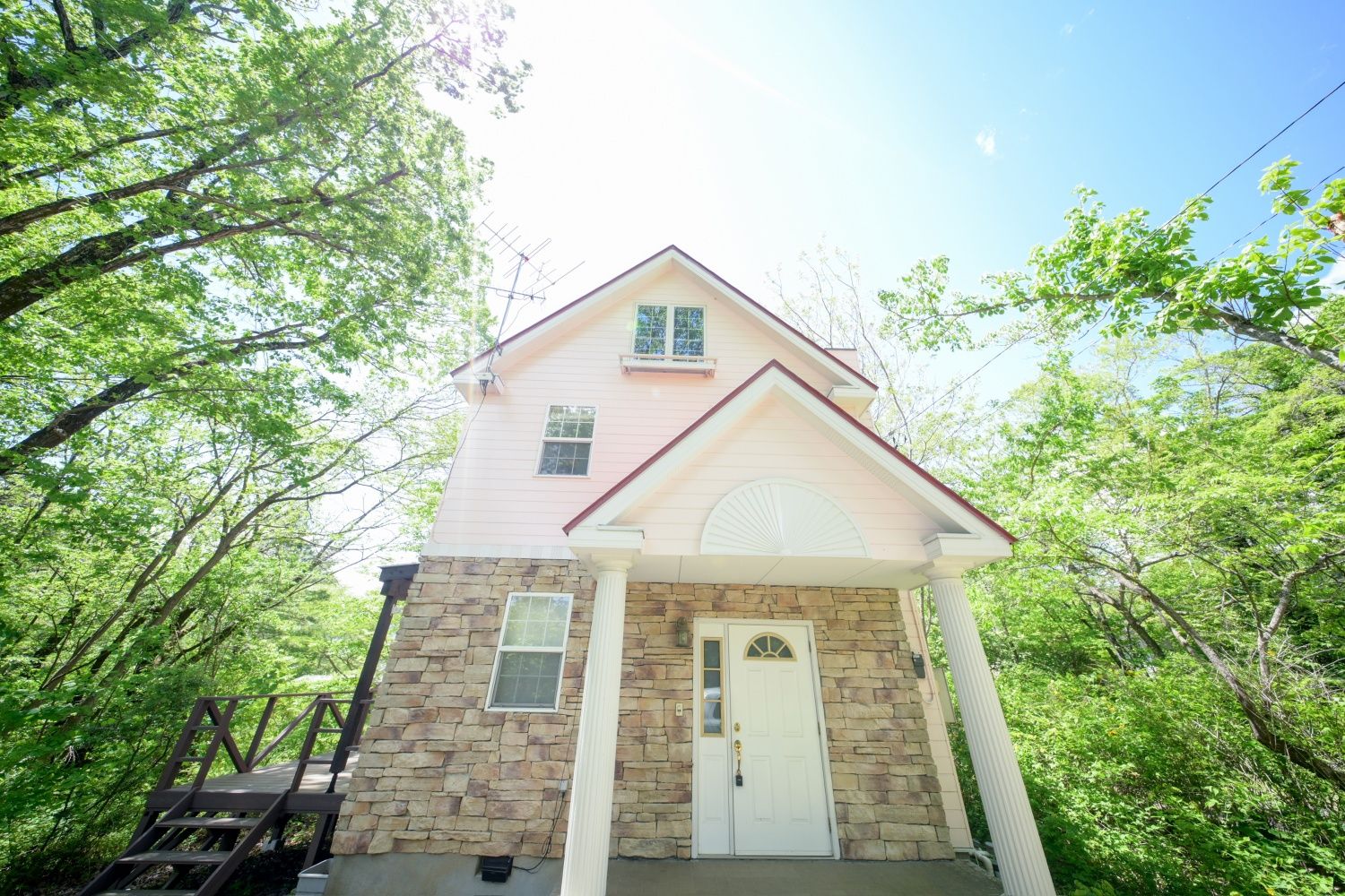 A two-story house with a stone foundation and a pink upper story, surrounded by lush greenery.