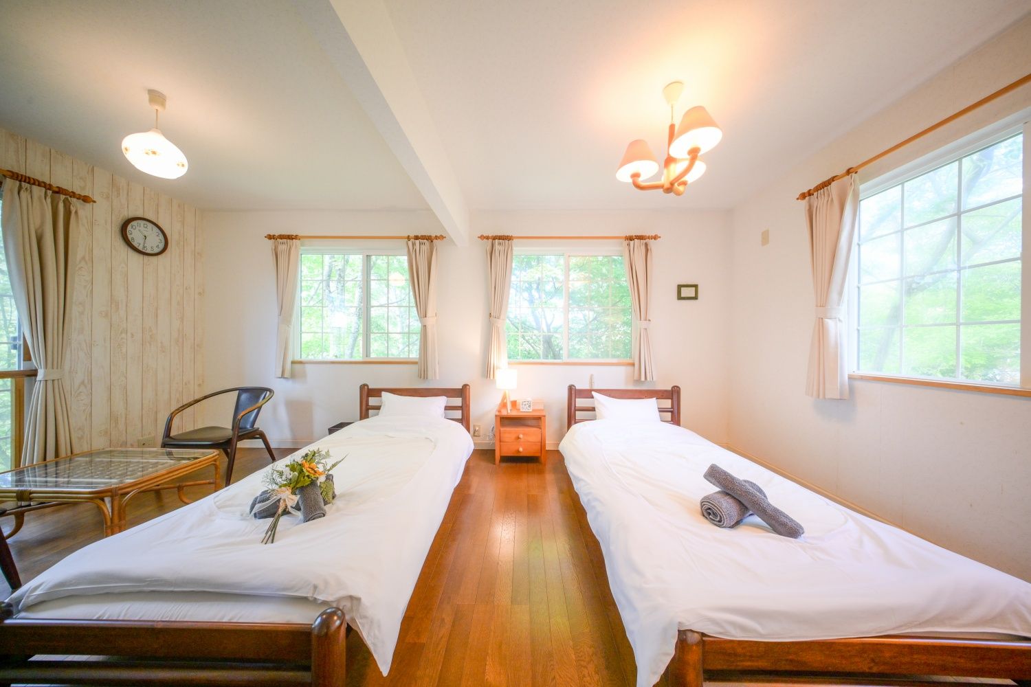 A room with two single beds, a wooden table with chairs, a clock on the wall, and a window with curtains.