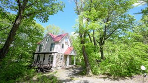 A two-story house with a stone foundation and a red roof, surrounded by dense green trees. - Yatsugatake Angel AG1 (Hokuto)