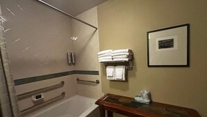 Um banheiro com banheira, chuveiro, porta-toalhas com toalhas dobradas, um quadro na parede e um banco de madeira com toalha e uma garrafa.