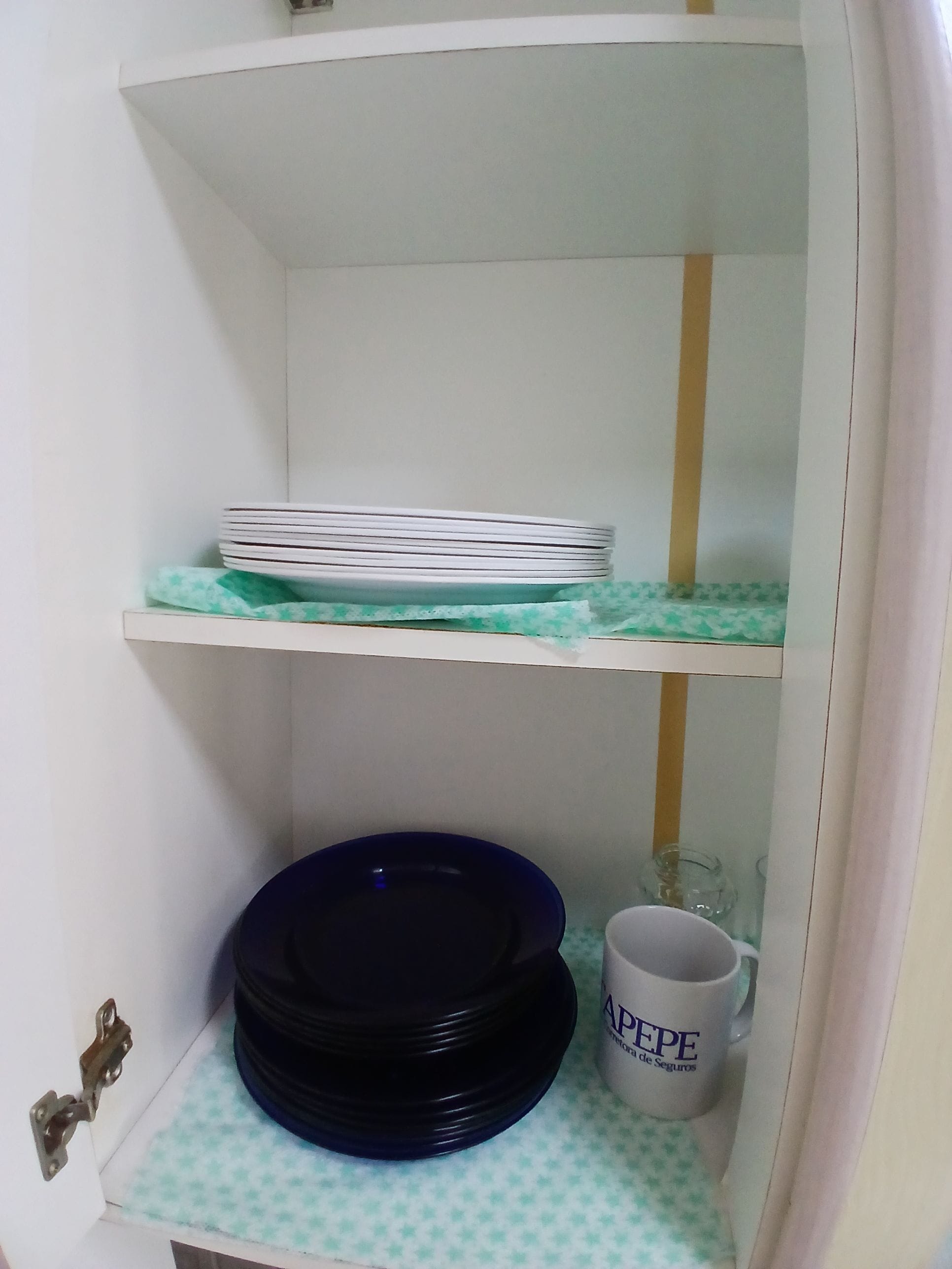 A white cabinet with two shelves; the top shelf holds stacked plates, and the bottom shelf has a blue bowl and a mug.