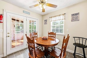 Un coin repas comprenant une table ronde en bois et quatre chaises, un ventilateur de plafond et une fenêtre avec des stores.