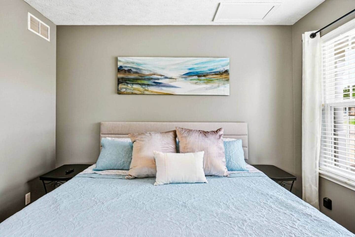 Un dormitorio con cama, mesitas de noche, un cuadro y una ventana con persianas.