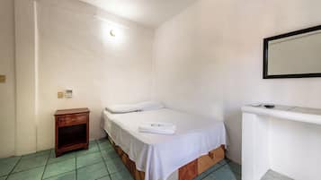 Pequeña habitación con una cama individual, una mesita de noche de madera, una colcha blanca y una pequeña mesa con una toalla.