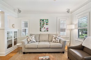 Ein Wohnzimmer mit einem beigen Sofa, zwei Zierkissen, einem Glastisch und einem Bücherregal.