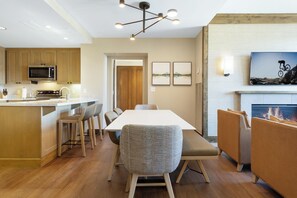 A modern kitchen with a central island, dining area with a long table and chairs, and a fireplace with a mounted TV. - Charlie Rentz: Remodeled, 2 Balconies, Ski-In/Out w/ Valet, Resort Amenities, Pool + Hot Tubs (Avon)