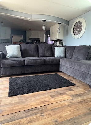 A grey sectional sofa with patterned cushions, a black rug, and a wooden floor. - Fabulous 3-bedroom mobile home with AC unit in Eagle Pass (Eagle Pass)