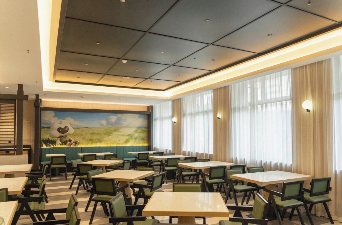 A hotel dining area with multiple tables and chairs, large windows with curtains, and a mural on the wall.