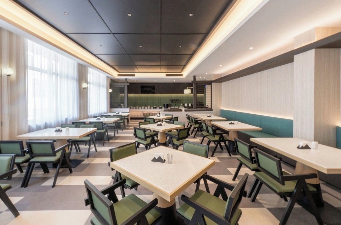 A modern dining area with multiple tables and chairs, a kitchen in the background, and large windows allowing natural light.