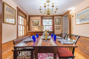 A dining room with a long wooden table set for six, surrounded by chairs with red cushions. A chandelier hangs from the ceiling, and the walls are adorned with framed pictures. - Brackett Cottage: Near Salem! Heart of Historic Marblehead; Walk to Shops & Dining (Marblehead)