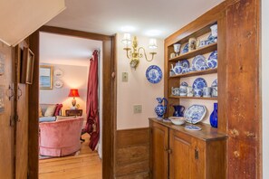 Un pasillo con un mueble de madera que exhibe objetos de cerámica azul y blanca, una silla roja y una lámpara.