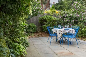 Un patio con mesa y sillas, rodeado de exuberante follaje y plantas con flores.