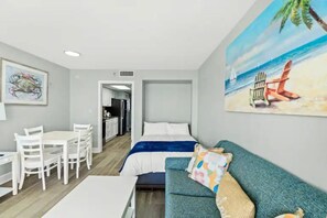 A room with a bed, a sofa, a dining table with chairs, and a painting of a beach scene. - Luxury Oceanfront 1BR | Designer Decor (North Myrtle Beach)