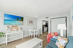 A modern living room with a dining area, a kitchen in the background, and a beach-themed painting on the wall. - Luxury Oceanfront 1BR | Designer Decor (North Myrtle Beach)