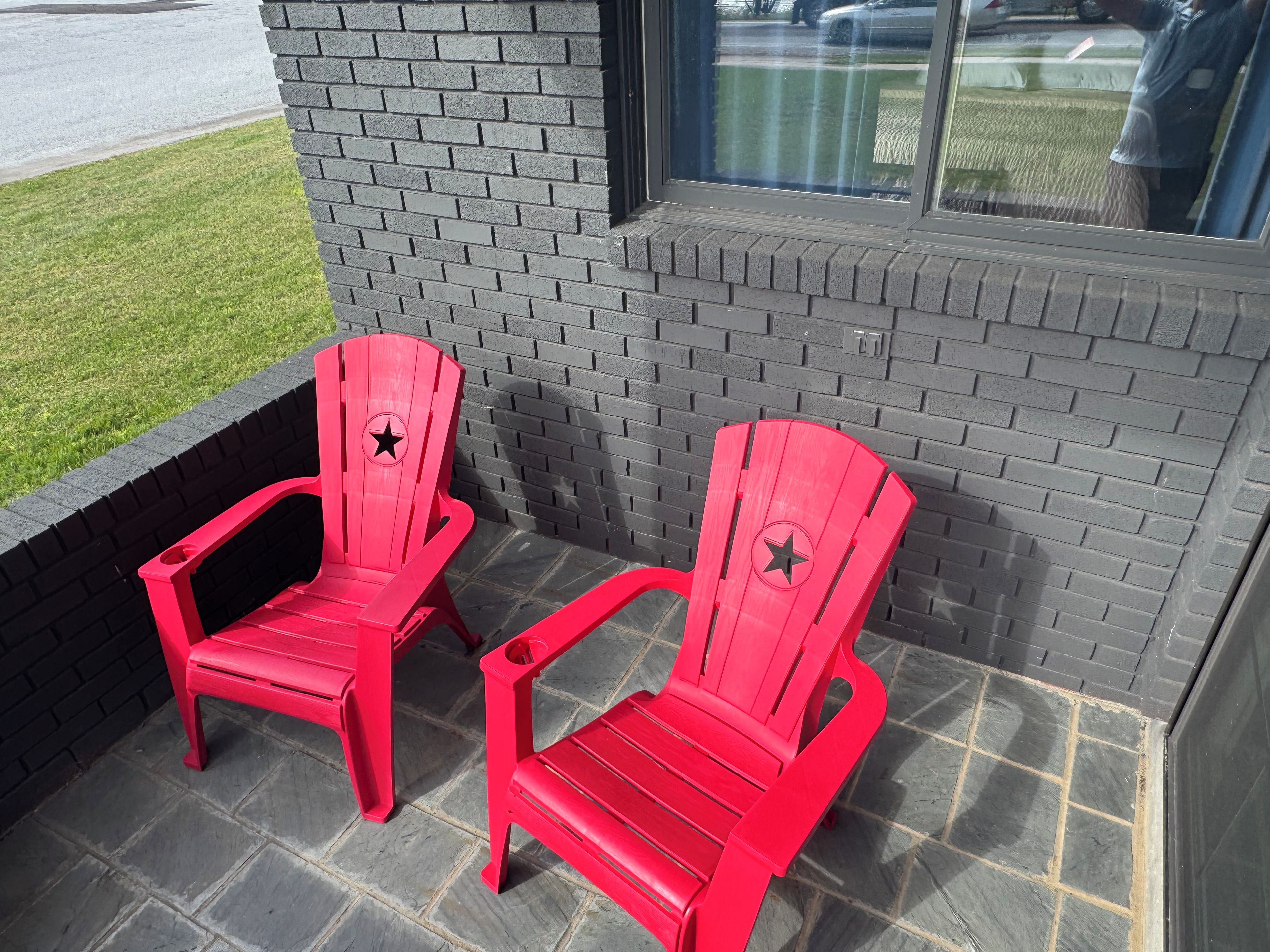 Two red Adirondack chairs on a stone patio with a brick wall and a window in the background.