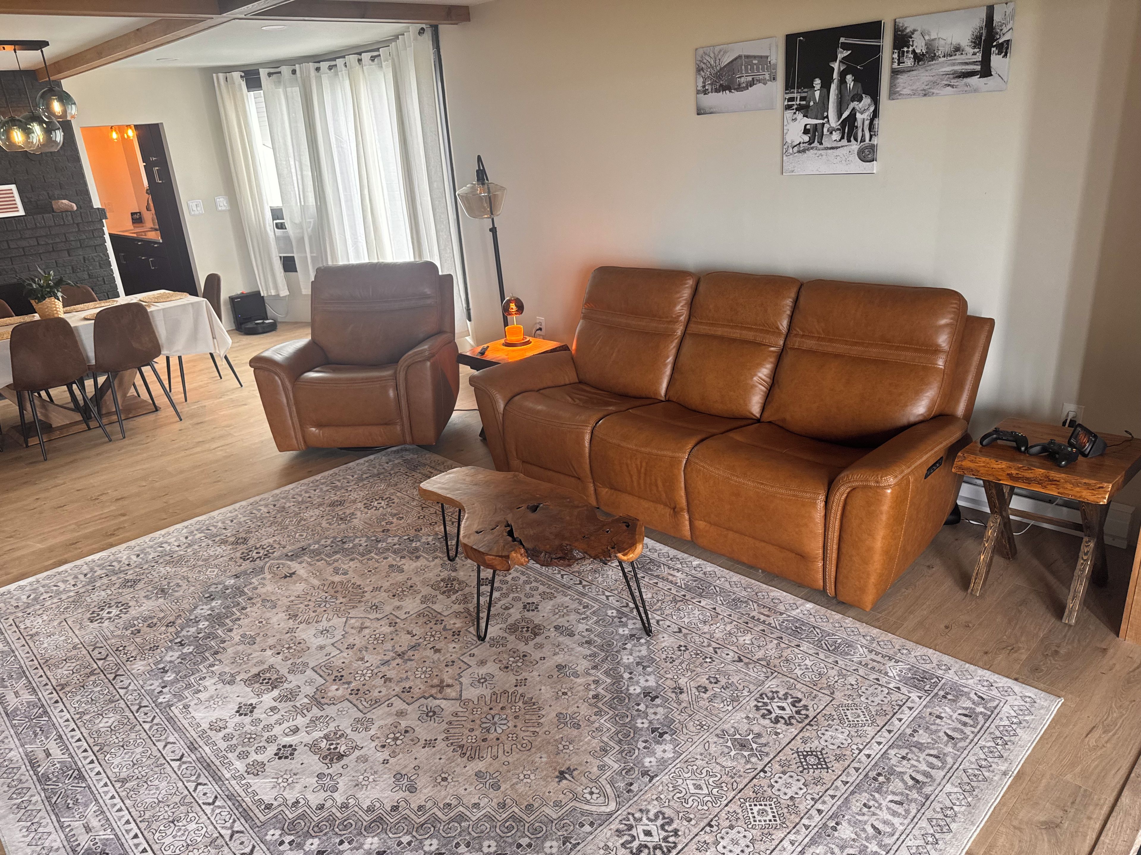 A living room with a brown leather sofa, a patterned rug, a dining area with a table and chairs, and a kitchen in the background.