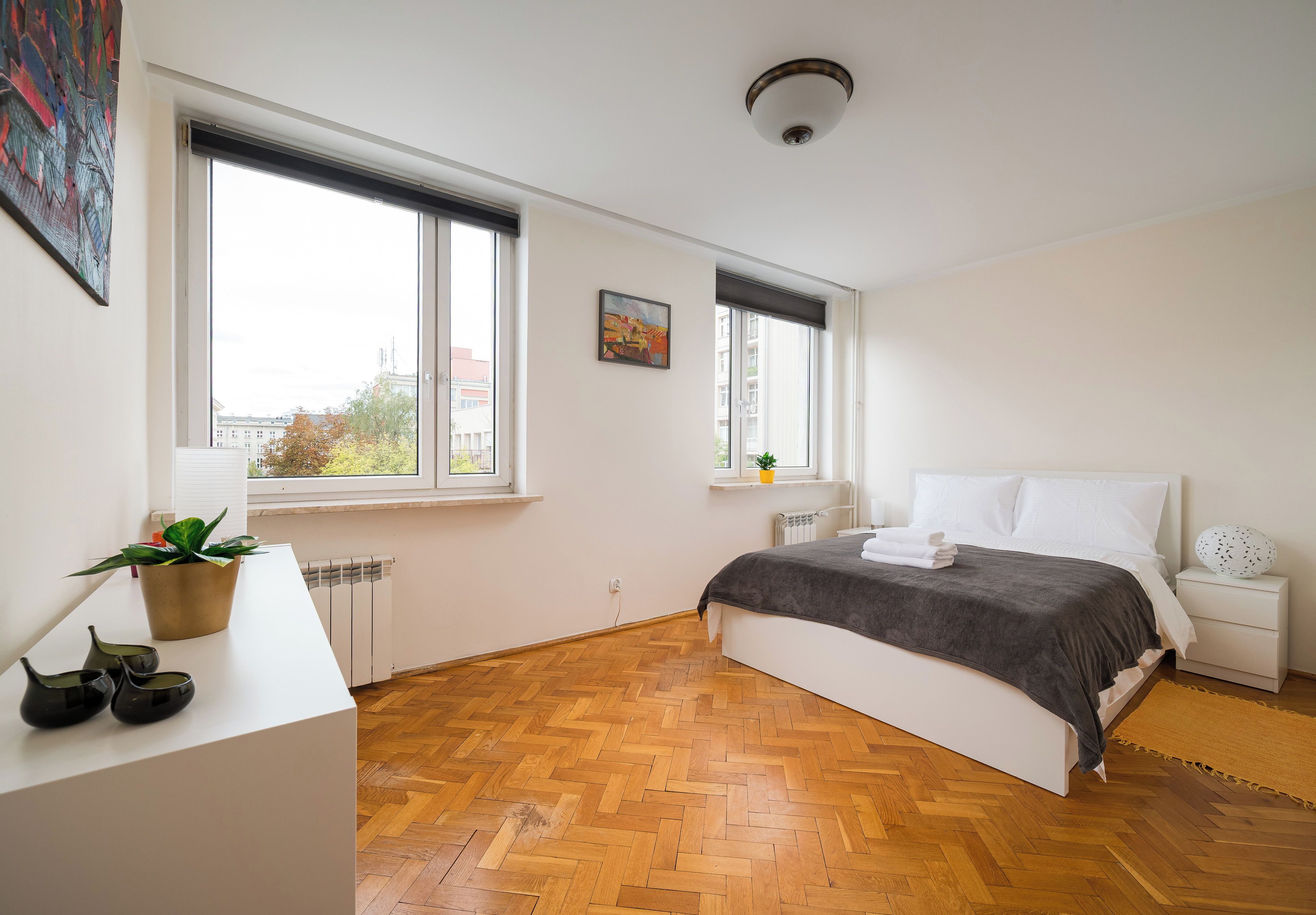 A bedroom with a bed, bedside tables, a radiator, and a window with a view of buildings.