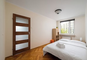 A bedroom with a wooden door, a radiator, a window with a view of buildings, and a bed with folded towels. - Warsaw Royal Route Apartment (Warszawa)