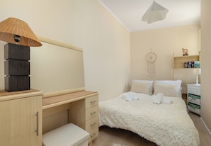 Ein Schlafzimmer mit Holztisch, einem Bett mit flauschiger weißer Bettwäsche, einem hängenden Traumfänger und einem Regal mit Büchern und einer Lampe.