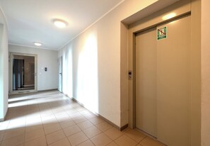 Een gang met een deur waarop een rookverbodssign staat, een digitaal bedieningspaneel van een lift en een glazen deur aan het einde.