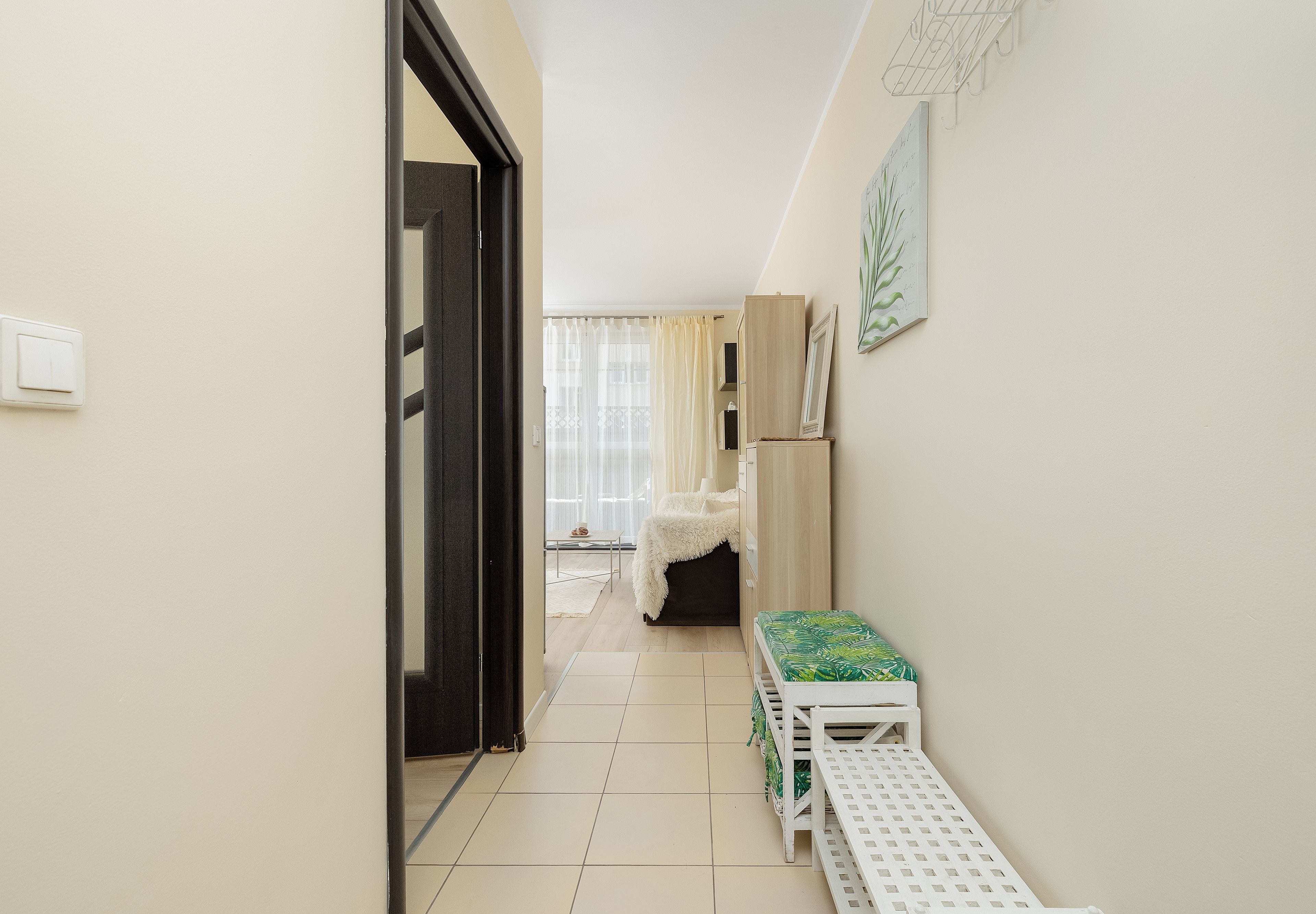 Un couloir étroit, avec une porte sur la gauche, un banc blanc orné d’un coussin à motifs sur la droite, et une fenêtre avec des rideaux légers en arrière-plan.