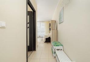 A narrow hallway with a door on the left, a white bench with a patterned cushion on the right, and a window with sheer curtains in the background. - Dunikowskiego Cosy Apartment (Szczecin)