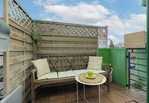Een houten zitgedeelte buiten met kussens en een geweven tafel, omringd door een houten hek en een groene wand.