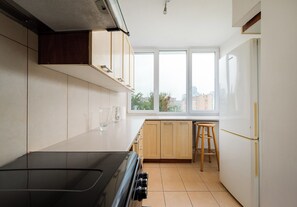 A kitchen with a black stove, wooden cabinets, a white refrigerator, and a window with a view of buildings. - Złota Spacious Apartment (Warszawa)